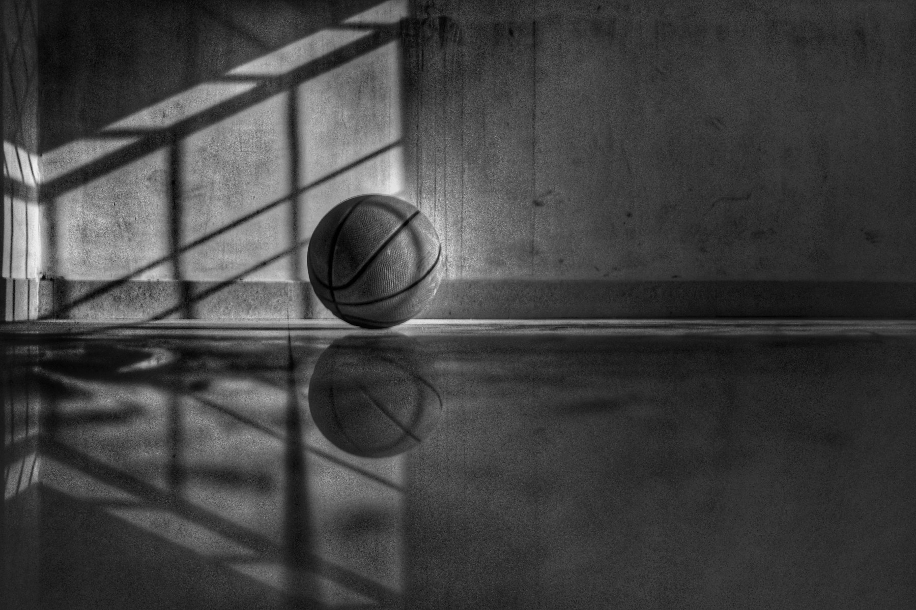 Black and white photograph of a basketball on a polished floor, in long architectural shadow.